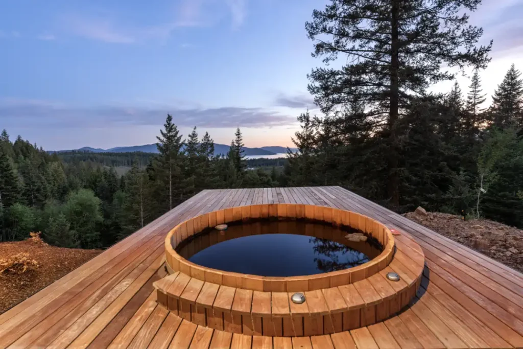 Round wood hot tub set into a deck at Basking Moose, with evergreen trees and Lake Pend Oreille in the distance at dusk.