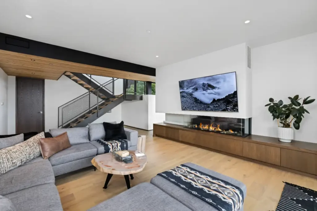 Lower-level living area at Basking Moose with a gray sectional, glass-fronted fireplace, wood cabinetry, and a steel staircase with wood treads and metal mesh railings.