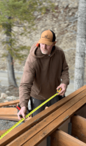 A builder wearing a Smithwright hat measures a deck board with a tape measure at a residential construction site.