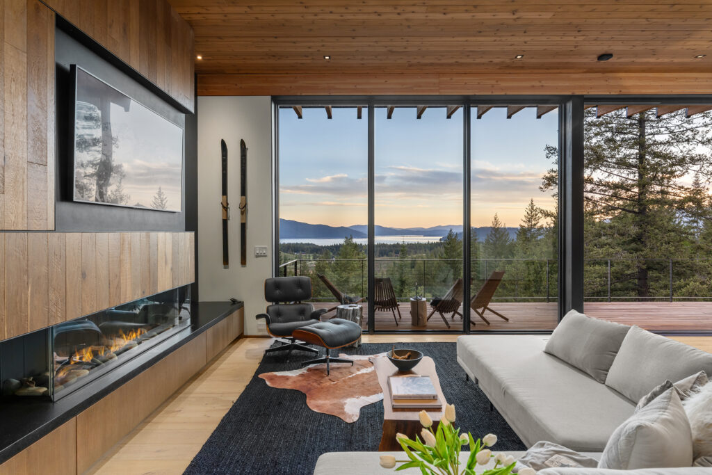 Living room at Basking Moose with a wood ceiling, glass-fronted fireplace, and floor-to-ceiling windows opening onto a deck overlooking Lake Pend Oreille.